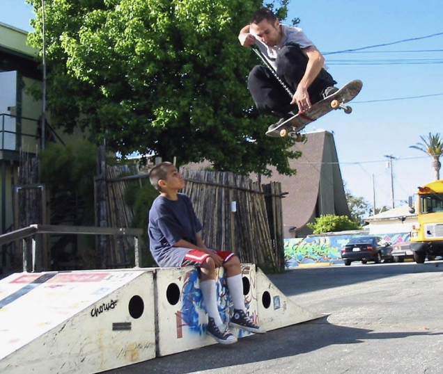 Skateboarder performing a high jump off a rap getting 7 feet of air with Sk8leash, jumping above another person with trees and buildings in the background.